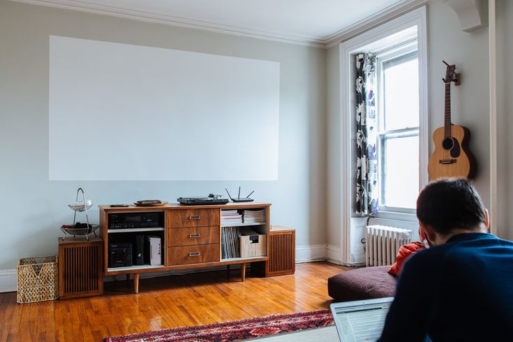 a man sitting in front of a tv on top of a hard wood floor next to a window