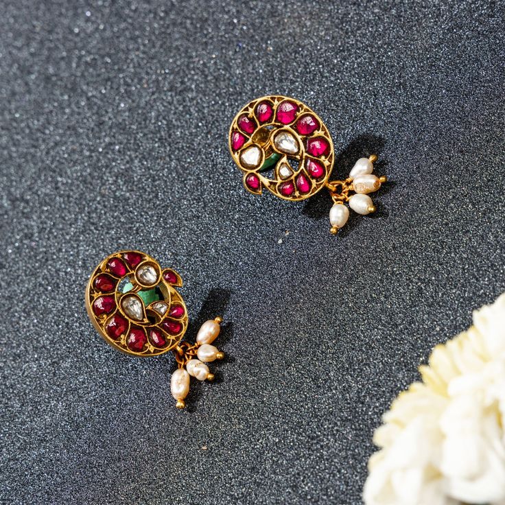 Introducing Neema Peacock Studs by Yamoona—a blend of heritage and elegance. The red ruby stud features a peacock shape with white stones. Delicate pearl hangings add femininity. Meticulous craftsmanship preserves tradition with a modern touch. The design symbolizes the regal peacock, deeply rooted in Indian culture. The combination of the red and white stones and pearls creates a timeless, elegant look. These studs effortlessly elevate your style for any occasion, leaving a lasting impression w Ruby And Pearl Earrings, Elegant Chandbalis With Peacock Design For Diwali, Festive Red Peacock Design Earrings, Elegant Chandbalis With Peacock Design For Celebration, Traditional Red Pearl Drop Earrings, Festive Red Peacock Earrings, Elegant Ruby Chandbalis With Meenakari, Red Peacock Design Earrings For Gifts, Red Peacock Design Earrings For Celebration