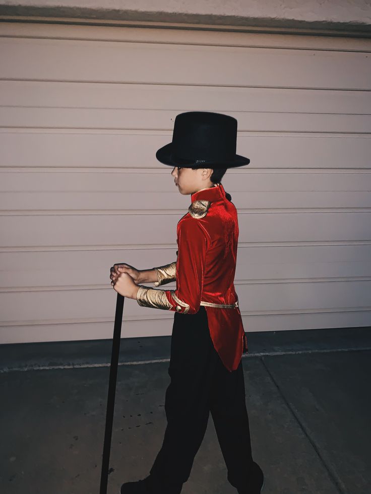 a young boy in a red jacket and black top hat holding a cane while walking towards a garage door