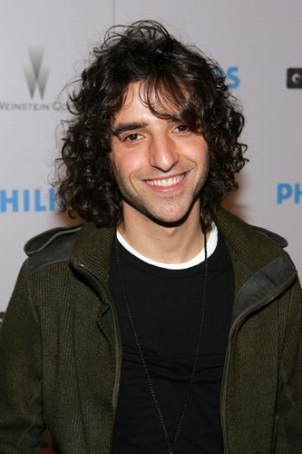 a man with long curly hair smiles at the camera while wearing a black shirt and green jacket