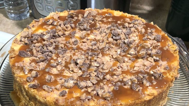 a cheesecake covered in caramel and nuts on top of a glass cake plate