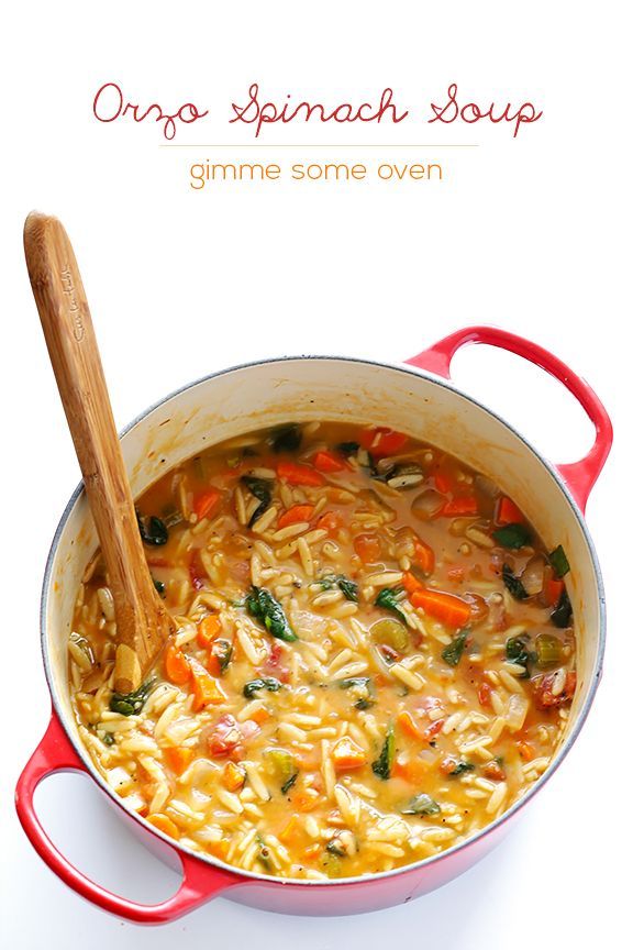 a red pot filled with pasta and vegetables on top of a white table next to a wooden spoon