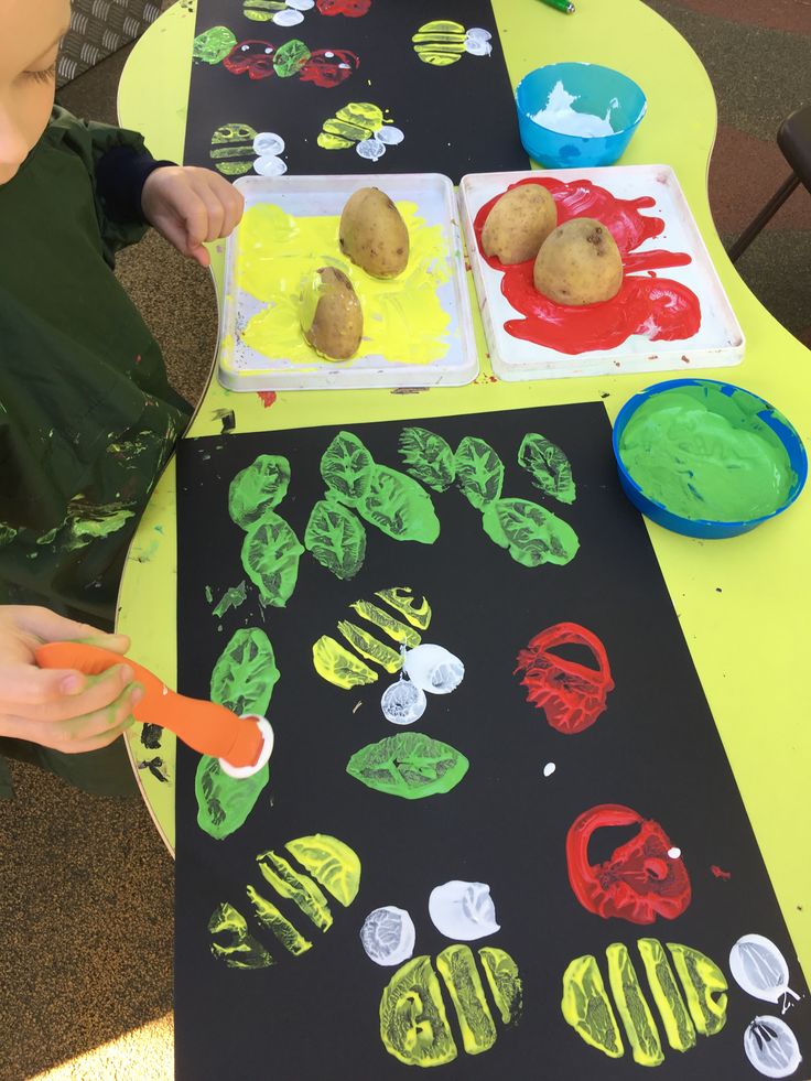 a young boy is painting potatoes on a black paper with green and red paint while sitting at a yellow table