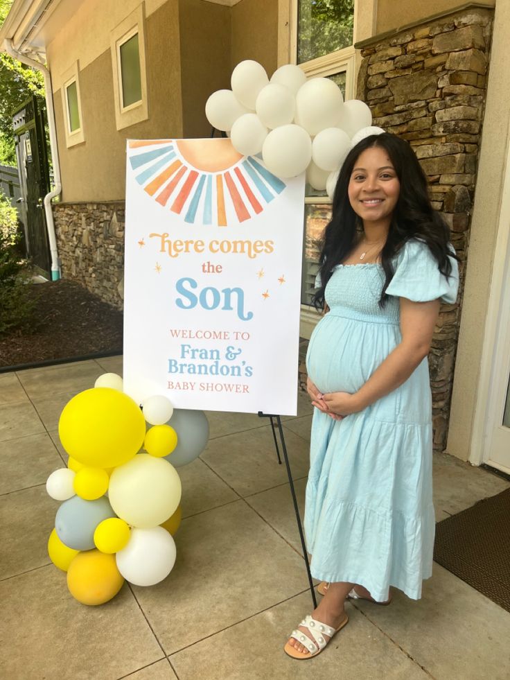 a pregnant woman standing in front of a sign for her new baby shower, with balloons all around her