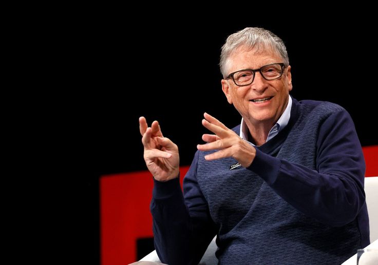 bill gates is sitting in a chair with his hands out and he is wearing glasses