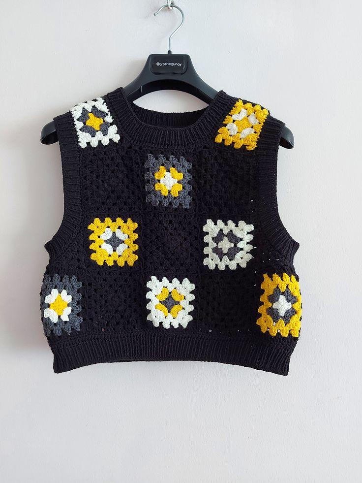 a black sweater with yellow and white crochet flowers on the front is hanging from a hanger