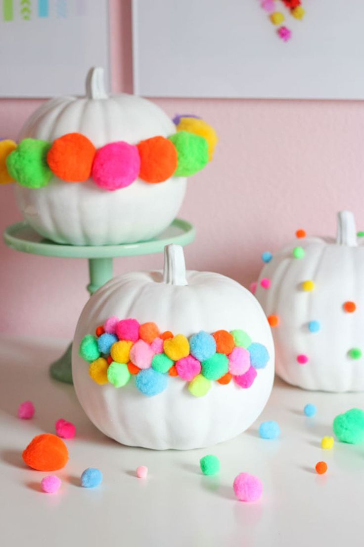 two white pumpkins decorated with colorful pom - poms on a cake stand