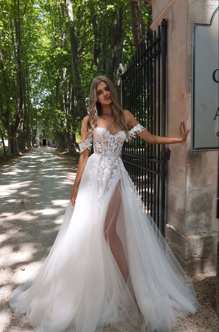 a woman standing in front of a gate wearing a white dress and high slit skirt