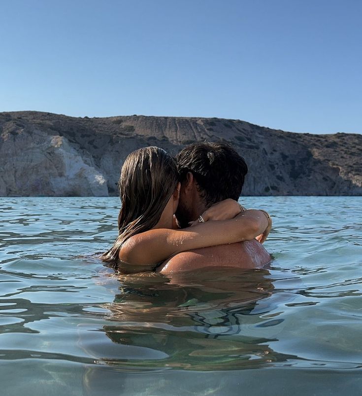 two people are in the water hugging each other and looking into the distance with mountains in the background