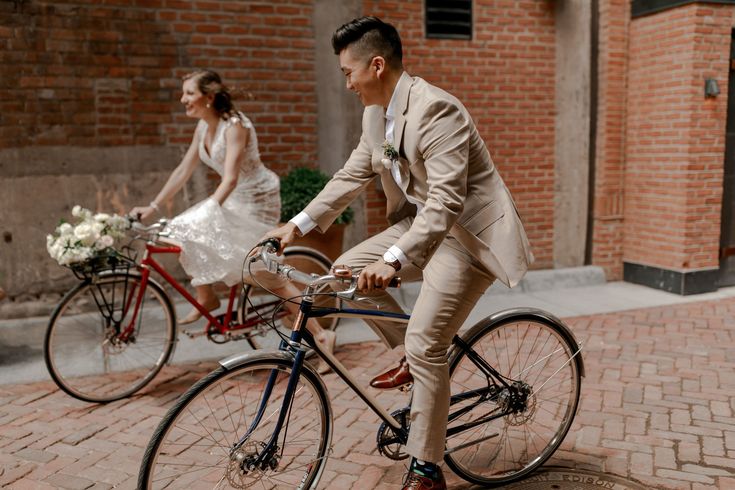 the bride and groom are riding their bikes down the street in front of brick buildings