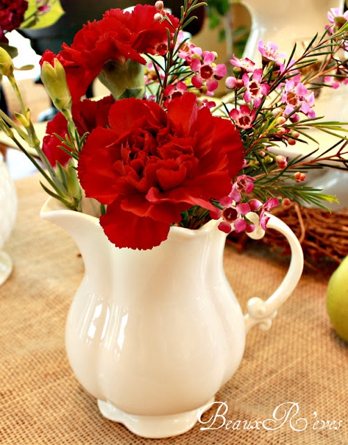 a white pitcher filled with red flowers on top of a table next to an apple