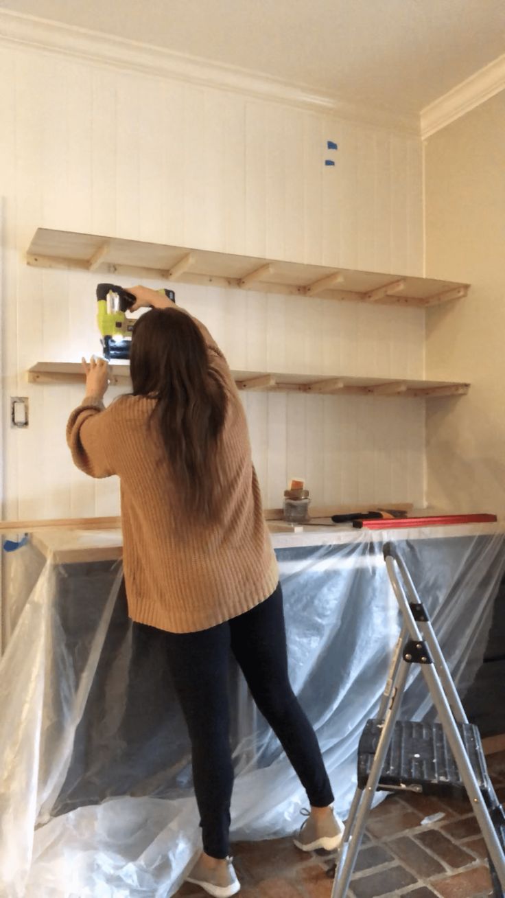 a woman is using a power drill to paint the wall in her kitchen with wood shelving