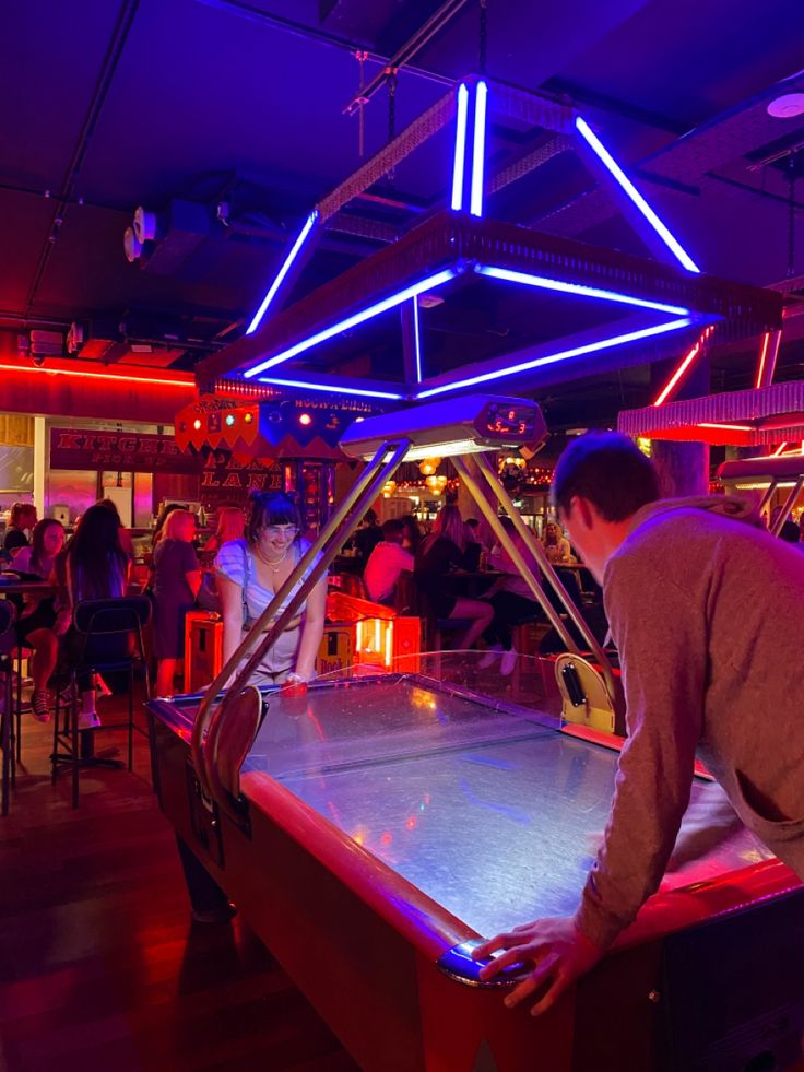 a retro arcade bar lit with neon lights 
and two people are playing air hockey Arcade Date Aesthetic Couple, Arcade Aesthetic Friends, Arcade Date Aesthetic, Retro Arcade Aesthetic, Arcade Pictures, Arcade Date, Date At Home, Fair Date, Arcade Aesthetic