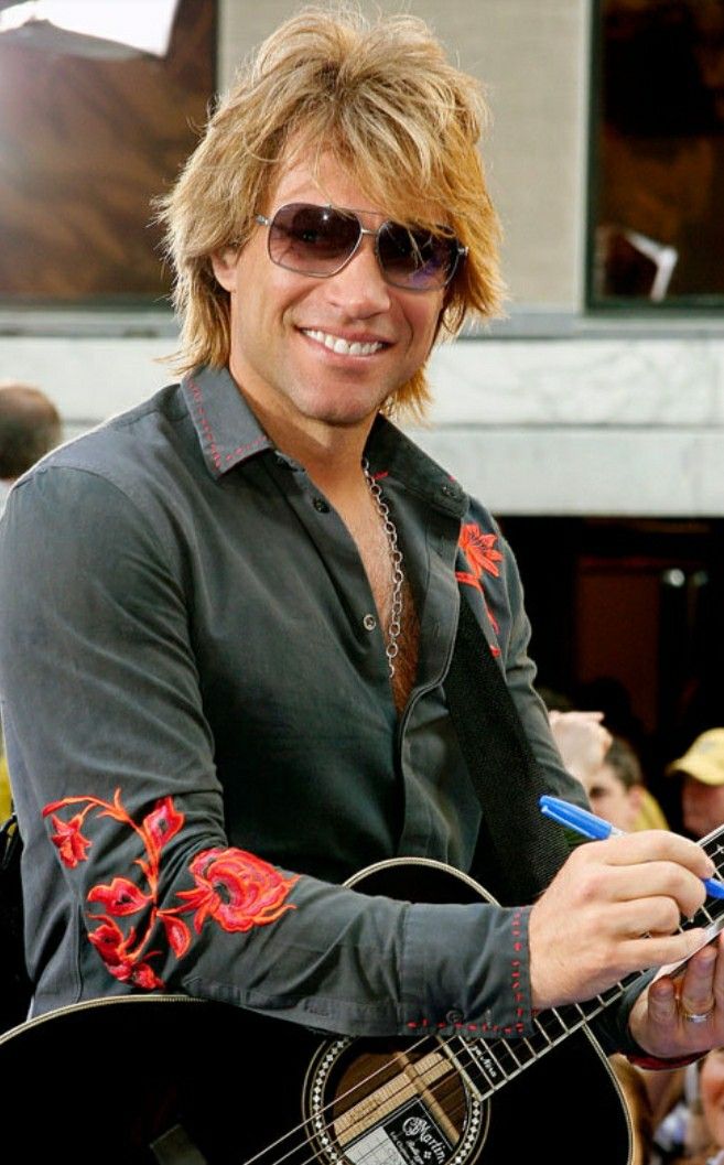 a man with sunglasses on holding a guitar and smiling at the camera while standing in front of a crowd