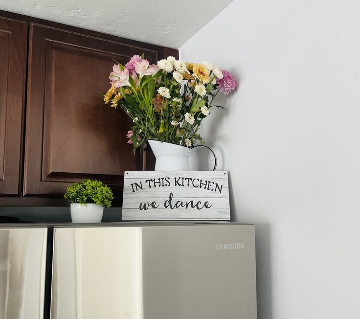 a vase with flowers and a sign that says in this kitchen we dance on top of a refrigerator