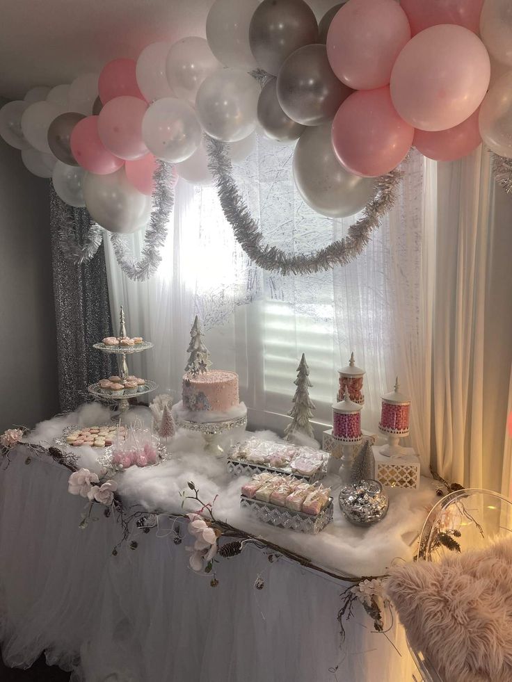 a table topped with lots of pink and silver balloons next to a window covered in snow