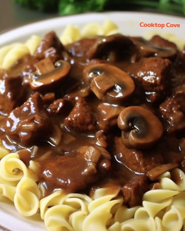 a white plate topped with pasta covered in meat and mushrooms on top of saucey sauce