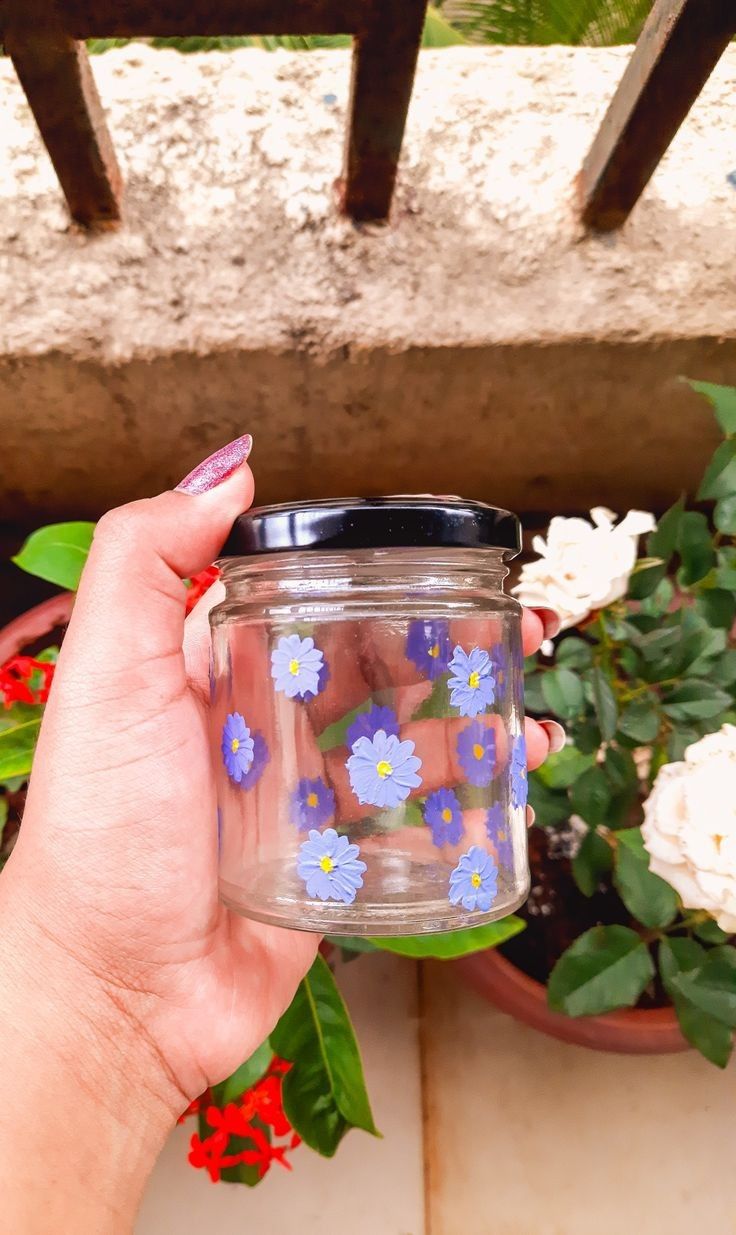 a hand holding a jar filled with blue and white flowers next to potted plants