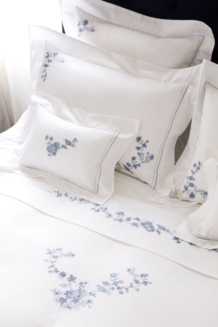 a bed with white sheets and blue embroidered flowers on the comforter, along with two pillows