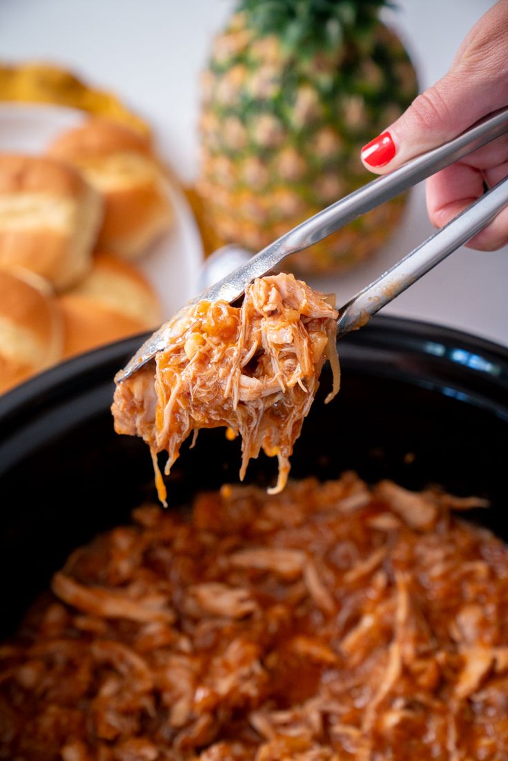 a spoon full of shredded chicken being lifted from a crock pot with pineapples in the background