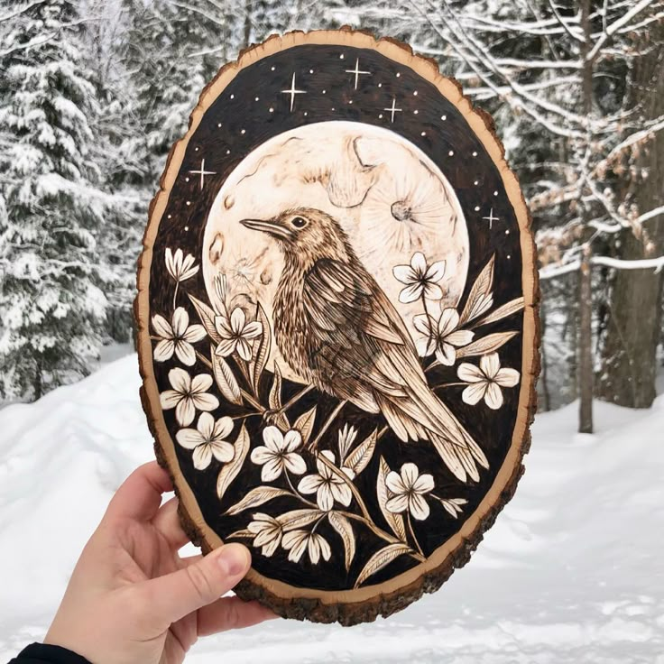 a hand holding up a wooden plaque with a bird on it in front of snow covered trees