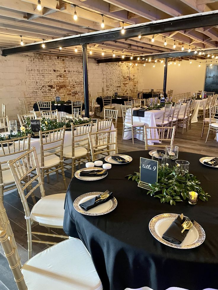 tables set up for an event with white chairs and black tablecloths, gold chargers and greenery