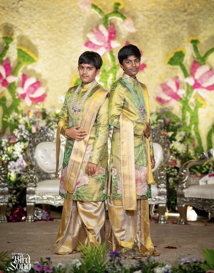 two young men standing next to each other in front of a floral wallpapered backdrop