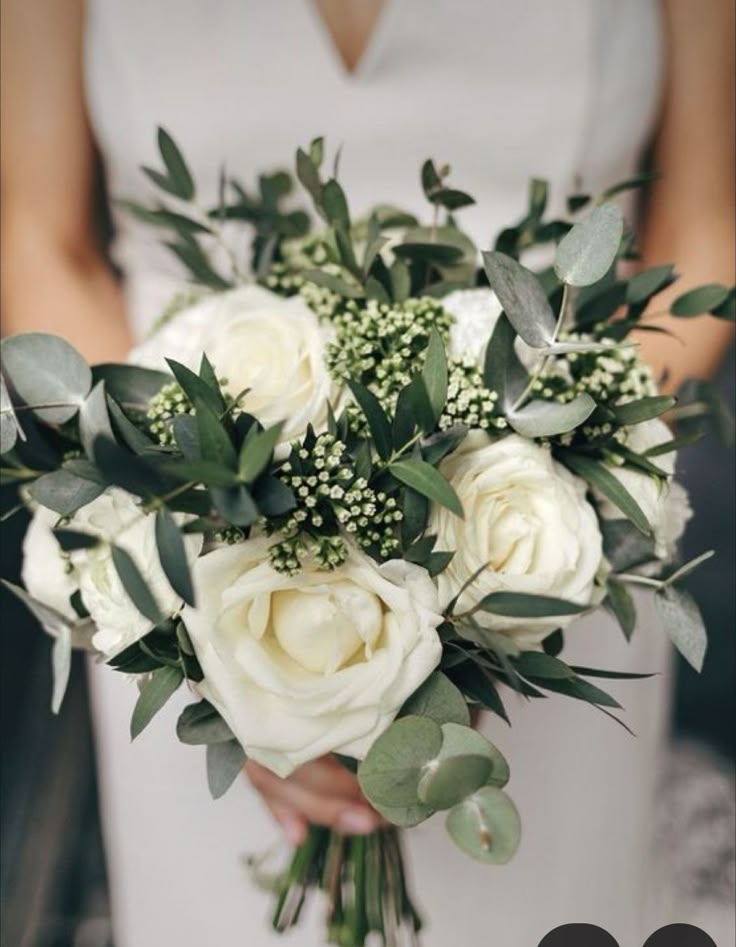 a bridal holding a bouquet of white roses and greenery in her hands,