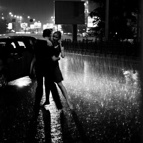 a man and woman hugging in the rain with their arms around each other as they stand next to a car