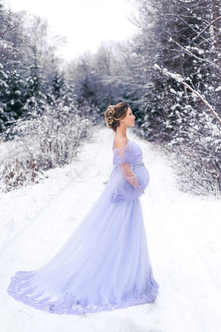 a pregnant woman in a blue gown standing on snow covered ground with trees behind her