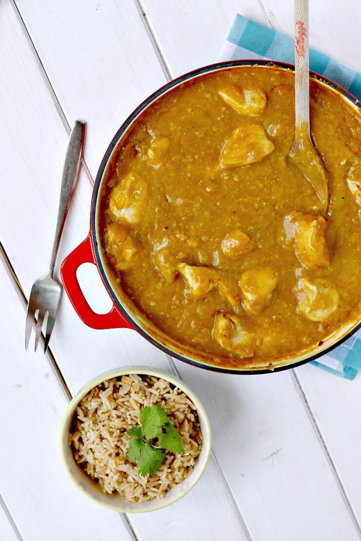 a pot of curry next to a bowl of rice and a spoon on a white table