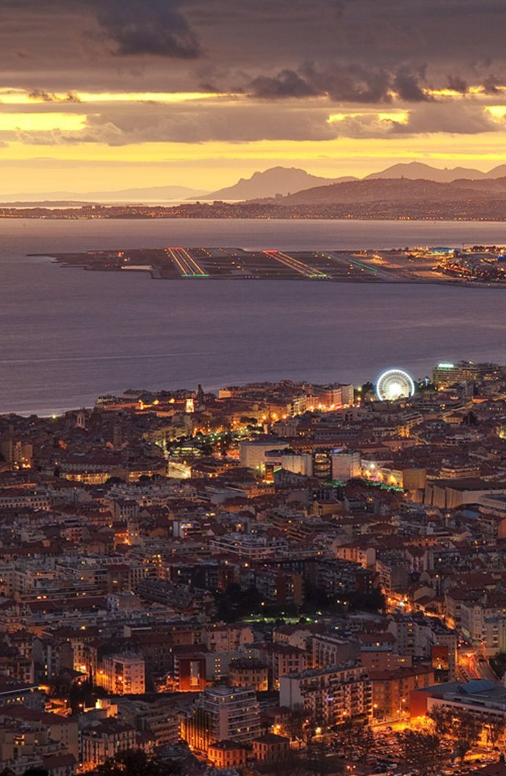 the city is lit up at night with mountains in the backgrouds and water