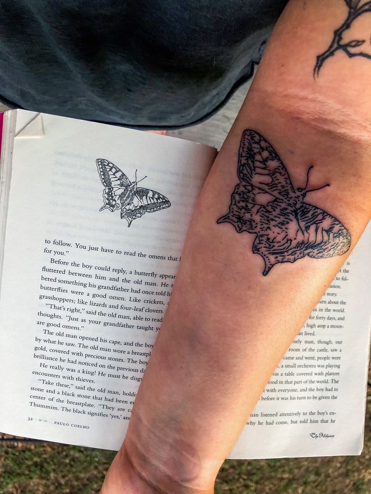 a person with a butterfly tattoo on their arm next to an open book that is sitting on the ground