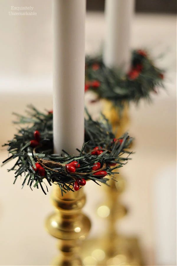 two white candles with red berries on them are sitting in a gold candle holder that is decorated with greenery and pine cones