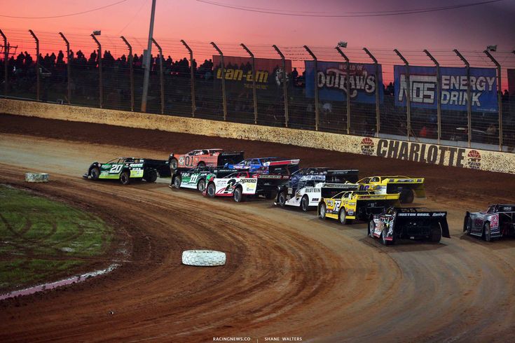 a group of dirt racing cars driving down a race track at sunset or sunrise with the sun setting in the background