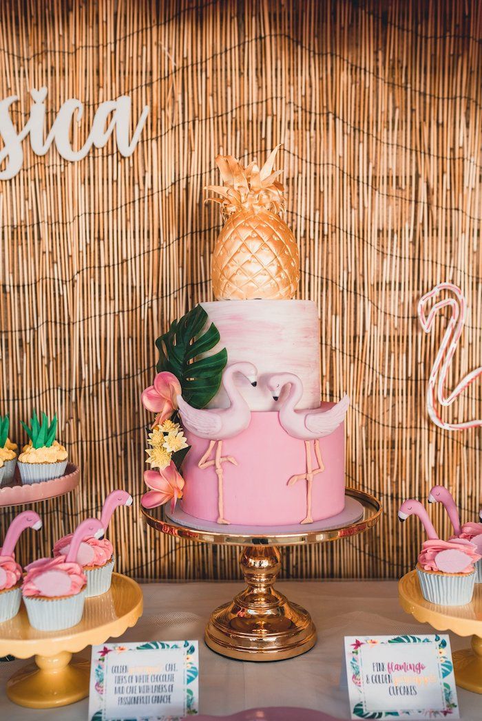 a pink cake with flamingos on top and cupcakes next to it in front of a bamboo wall