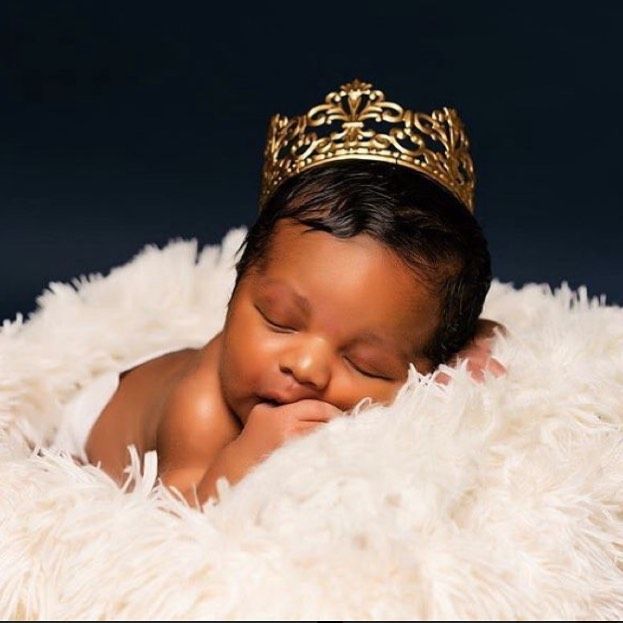 a baby wearing a golden crown laying on top of a white fur covered blanket with his hands under his chin