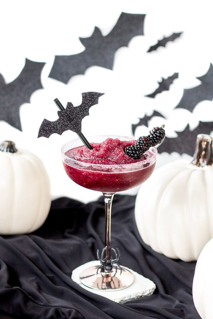 a halloween drink in a glass with berries and black bat decorations on the top, sitting next to white pumpkins