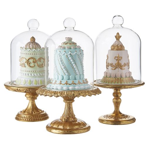 three cakes under glass covered with frosting on top of each other in gold stands