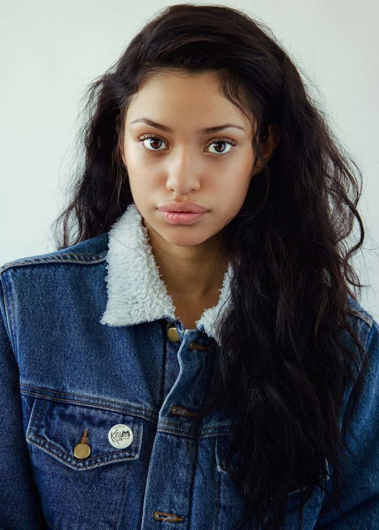 a young woman with long hair wearing a denim jacket and looking at the camera while standing in front of a white wall