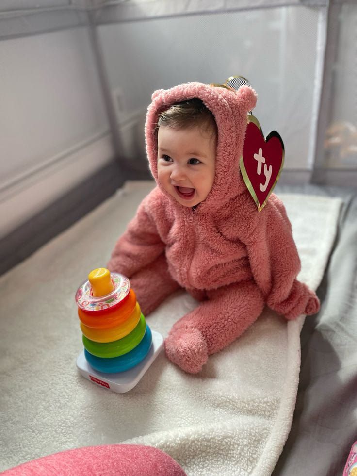 a baby in a pink teddy bear outfit sitting on a bed next to a stack of toys