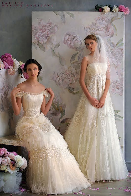 two women in wedding gowns standing next to each other with flowers on the wall behind them