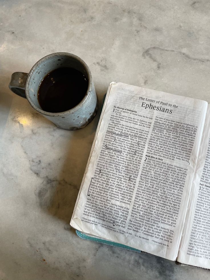 an open bible next to a cup of coffee on a marble counter top with a spoon