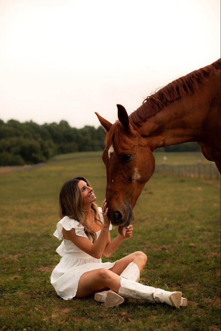 a woman sitting on the ground next to a brown horse in a field with grass