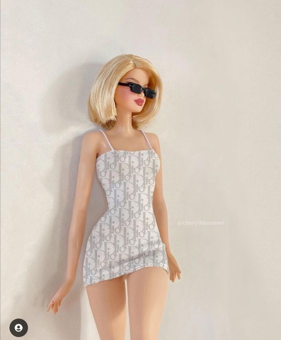 a doll is posed in a white dress and black sunglasses on her head, leaning against a wall