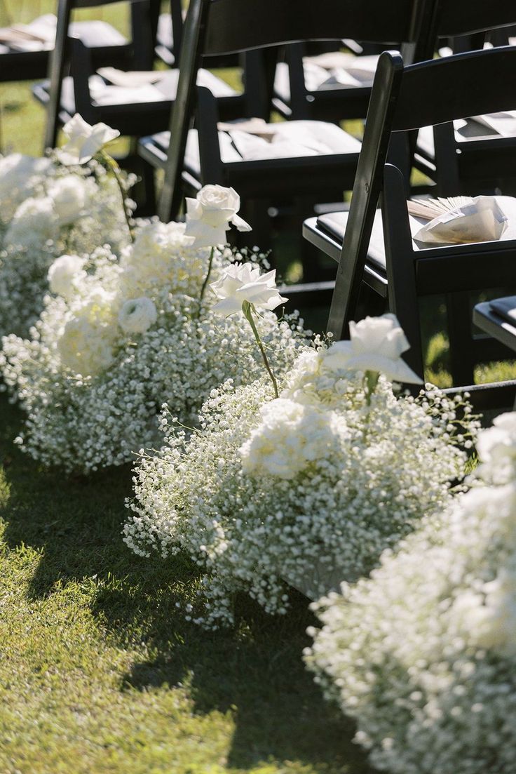 rows of black chairs with white flowers on them in the grass at an outdoor ceremony