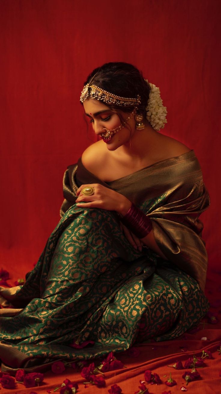 a woman in a green and gold sari sitting on a red background with rose petals