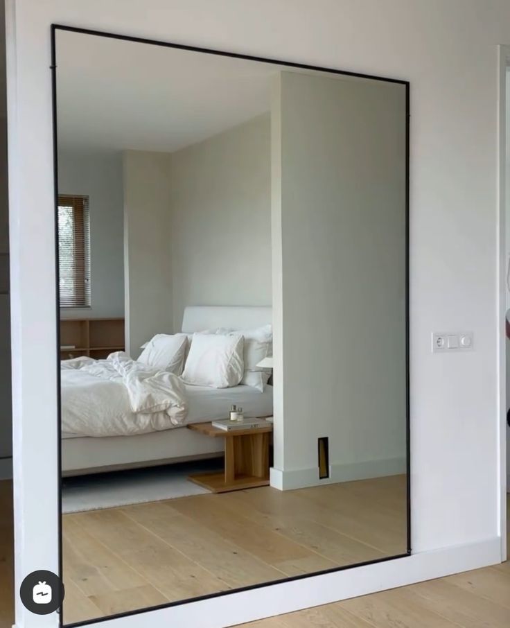 a large mirror reflecting a bed in a room with white walls and wood flooring