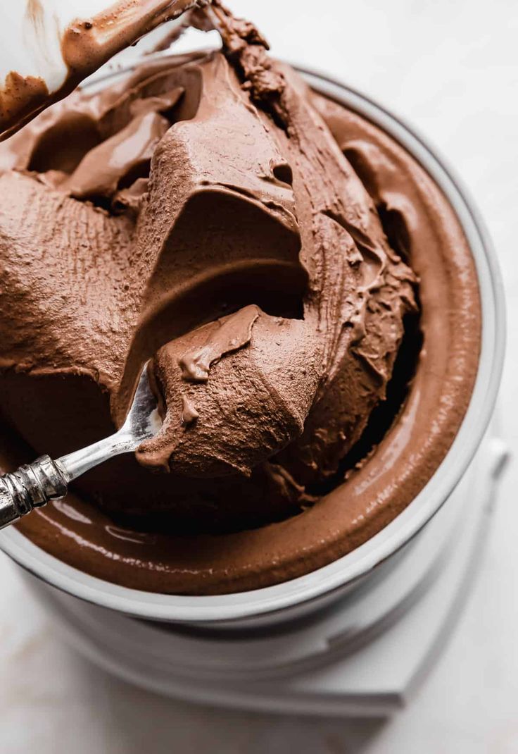 a bowl filled with chocolate frosting on top of a white table next to a spoon