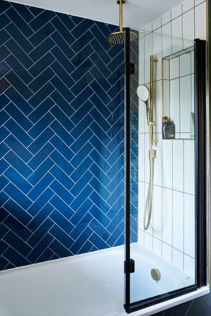 A close-up of a stylish shower area featuring navy blue herringbone tiles on the wall, contrasted by white vertical tiles on the adjacent wall. The shower is equipped with brass fixtures, including a rainfall shower head and a handheld shower. A sleek black-framed glass shower door adds a modern touch to the design, combining bold colours and luxurious accents for an elegant and contemporary look. Navy Herringbone Tile Bathroom, Shower With Brass Fixtures, Small Family Bathroom, Family Bathroom Design, Unique Bathrooms, Chevron Tile, Traditional Bathrooms, Modern Style Bathroom, Cage Light